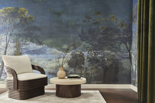 A living room with a dark wooden brown single seater with an off-white cushion, accompanied by a round side table in matching shades. A ceramic vase with decorative branches sits on the table, alongside a pile of books. The room features a dark wooden floor and an olive-colored velvet curtain on the right. The backdrop showcases a night landscape with trees from Omexco's Café Society wallcovering collection in shades of blue with green details. Delicately printed on fine linen, this panoramic design evokes the grandeur of the past while embracing the essence of contemporary design.