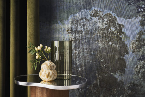 A decor shot featuring an olive-colored velvet curtain on the left. Next to it, an organically shaped side table holds a green vase and a smaller ceramic vase. The backdrop showcases a night landscape with trees from Omexco's Café Society wallcovering collection in shades of blue. Delicately printed on fine linen, this panoramic design evokes the grandeur of the past while embracing the essence of contemporary design.