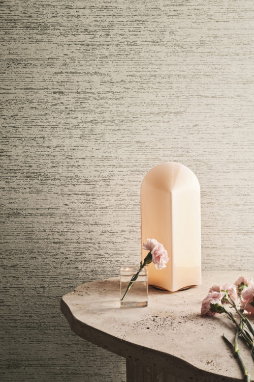 A close-up decor shot of an organically shaped stone table featuring a sleek table lamp and fresh cut pink flowers. The backdrop displays Omexco's textile wallcoverings from the Café Society collection in shades of off-white and light gray. This harmonious blend of distinct soft textures and colors creates visual depth and a vibrant rhythm.