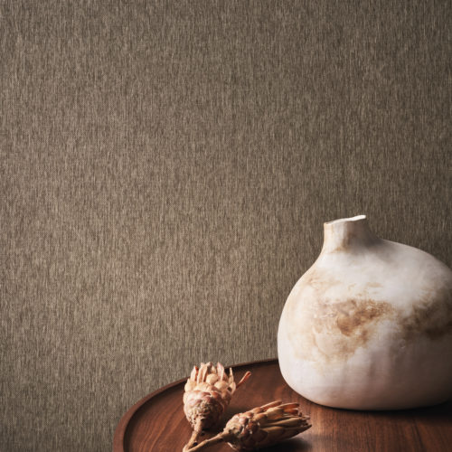 A close-up of a round wooden side table with an organic-shaped vase and two dried flowers on top. On the wall, embossed textile wallcoverings in chocolate brown from Omexco's Café Society collection are visible. This timeless classic with a glossy flair infuses spaces with a luminous glow.