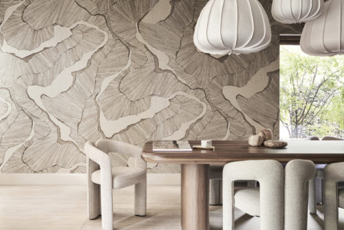 A stylish living room featuring a large wooden dining table surrounded by comfortable soft beige chairs. Above the table hang three large white pendant lights, providing a modern touch. On the left, a window offers a view of a garden with trees, bringing a sense of tranquility to the space. The wall is adorned with a panoramic design from Omexco's Café Society collection in shades of very light brown. The design, digitally printed on fine linen, makes a bold impression with its depiction of organic structures.