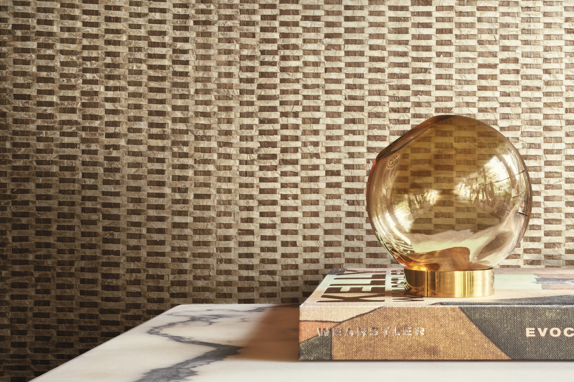 An elegant round glass table lamp, beautifully positioned on top of a book, serves as the focal point in this stylish decor. The backdrop features luxurious capiz wallcoverings from Omexco's Archipelago collection, showcasing a captivating blend of gold and brown shades, creating a sophisticated and inviting atmosphere.