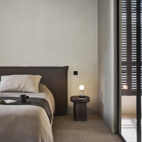 A tranquil bedroom in soothing shades of beige, featuring a glass sliding door separating the sleeping area from the bathroom. A plush, solid light brown carpet enhances the serene atmosphere. The double bed is adorned with an open book and a mug, inviting relaxation. The walls are adorned with Omexco's Silence wallcoverings in a calming beige hue, not only adding aesthetic appeal but also offering acoustic properties for a peaceful and quiet retreat