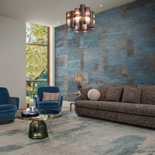 An image of a living room with bold, textured wallcoverings in shades of blue, which draws the eye towards the focal point of the room. These natural wallcoverings by Omexco have a leather-like look, but are actually made of Mulberry bark. The seating consists of a comfortable grey sofa and two stylish blue armchairs. The room is well-lit with natural light from a large window and a ceiling light fixture. Decorative elements such as a large rug in blue tones, a round coffee table in green glass and an organic shaped table lamp add interest and balance to the space.