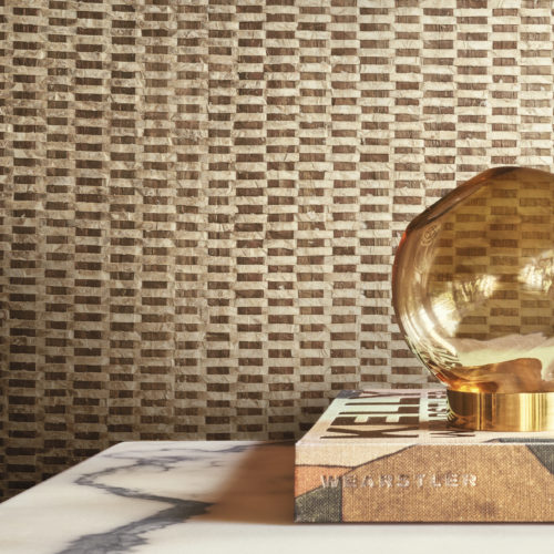 An elegant round glass table lamp, beautifully positioned on top of a book, serves as the focal point in this stylish decor. The backdrop features luxurious capiz wallcoverings from Omexco's Archipelago collection, showcasing a captivating blend of gold and brown shades, creating a sophisticated and inviting atmosphere.