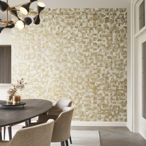 A sophisticated dining room featuring an oval dining table surrounded by soft beige chairs atop a white carpet, complemented by a herringbone parquet floor. The room is illuminated by a striking black and gold hanging lamp. The focal point is the captivating Omexco wallcovering collection, Archipelago, showcasing a patchwork of meticulously selected capiz pieces in shades of gold and beige. The textured wallcovering adds a luxurious and eye-catching element to the overall design of the room.