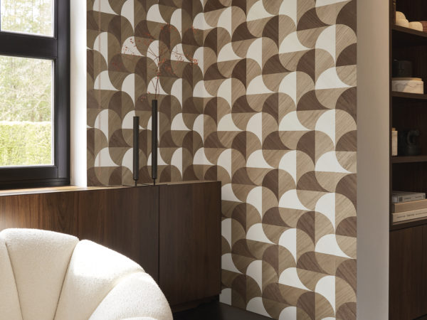 A cozy living room corner featuring a dark grey parquet floor, a white comfortable couch on the left and a window, providing natural light. On the right, built-in wall shelves display books. Adorning the wall is Omexco's marquetry collection Imperialis, showcasing geometric shapes in light brown, dark brown, and white, inspired by the graceful shape of lotus leaves.