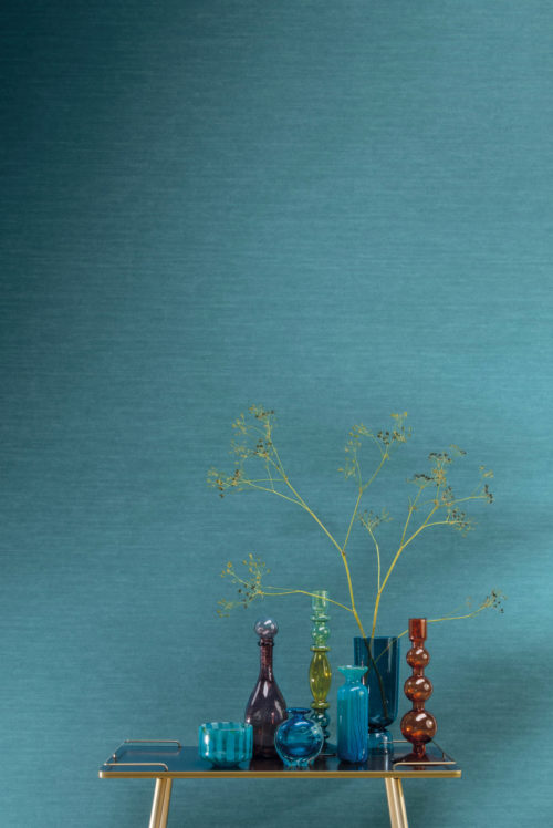 Closeup of a vivid turquoise wall. This is Omexco’s most popular signature plain, printed on a textured non-woven to give it that familiar textile look. The design of this wallcovering is called «Silky» and belongs to the tropical «Birds of Paradise» collection. In front of the wall we see a gold side table on which colourful glass vases of various heights and shapes are posed.