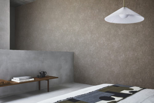 A room with a minimalist aesthetic. The back wall features a textured chocolate brown printed non-woven wallcovering from Omexco’s Edition collection. In the center, a hanging lamp with a white pleated lampshade is attached to the ceiling. To the left, a wooden bench holding books and a black teapot is placed in front of a smooth concrete wall. A bed is partially visible in the foreground, covered with a blanket featuring geometric patterns in black, gray, olive green, and beige. The floor is light-colored, complementing the modern and clean look of the room.
