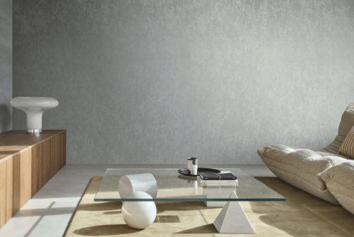 A minimalist living room with a plaster-like printed non-woven wallcovering from Omexco's "Edition" collection. The wall has a textured grey appearance. The room features a low wooden cabinet with a white lamp on top, a glass coffee table with white geometric decorative objects, and a beige cushioned sofa with a striped pillow. The floor has a light-colored rug, complementing the modern and clean look of the space.