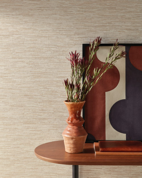 An image of a wooden side table featuring a brown ceramic vase filled with dried flowers, along with two brown notebooks and a painting of geometric shapes in rust, white and navy. The table is positioned against a wall on which Omexco’s Loom Stories wallcoverings are installed and is illuminated by natural light from a nearby window. This elegant linen and cotton weave is shown here in a soothing shades of beige and light brown. The combination of organic and geometric elements creates an interesting visual contrast, while the warm brown color scheme creates a cozy and welcoming ambiance.