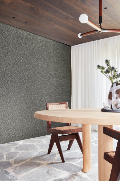 Image of a bright dining room with floor-to-ceiling white sheer curtains, a natural stone floor, a minimalist Scandinavian-style dining table with matching chairs and textured black and white Omexco wallcoverings. This linen bouclé yarn from the Loom Stories collection appears as stimulating to the eye as it does to the touch. The room is well-lit and spacious, with ample natural light streaming in through the windows. The simple yet elegant decor elements create a cozy and inviting atmosphere.