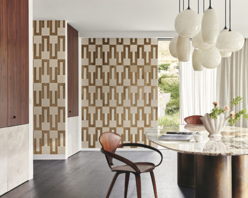 Image of a luminous living room with a dark parquet floor, featuring a large oval marble dining table supported by cylindrical legs in antiqued brass. A wood mid-century modern chair complements the table. In the background, a large window with a white curtain allows natural light to fill the space. Pendant lights with various lampshades in white glass illuminate the dining table. On the walls, Omexco's geometric marquetry collection "Imperialis" is displayed. It showcases a delicate interlocking pattern of wood against a metallic background. The shapes are dark brown, light brown and white, creating a sophisticated and harmonious atmosphere.