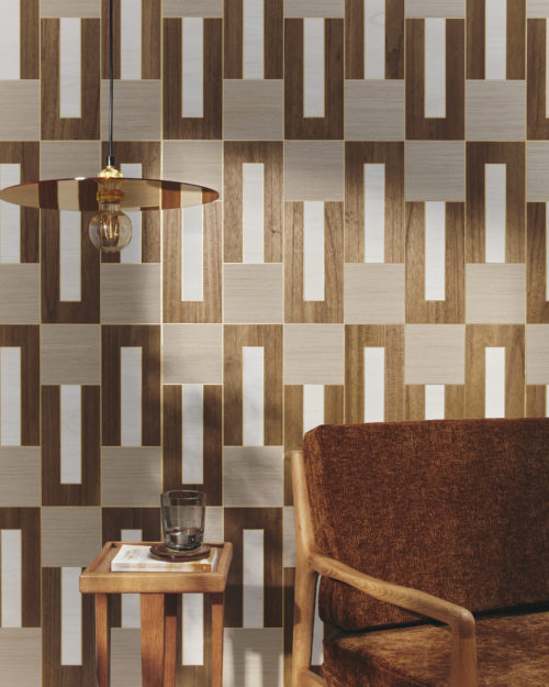 A close-up of a mid-century modern armchair with a wooden side table, adorned with a book and a glass. A sleek, minimalist hanging lamp, showcasing a visible lightbulb, illuminates the scene. Natural daylight gently graces the wall, on which Omexco's marquetry wallcovering collection "Imperialis" is displayed. It showcases a delicate interlocking pattern of wood against a metallic background. The shapes are dark brown, light brown and white, creating a sophisticated and harmonious atmosphere.
