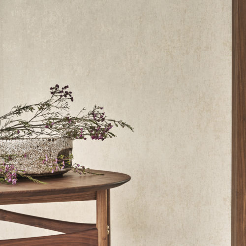 A minimalist interior featuring a printed non-woven wallcovering from Omexco's "Edition" collection. The wall has a textured beige appearance. In the foreground, a wooden table holds a speckled ceramic bowl filled with delicate branches and small purple flowers. The overall aesthetic is natural and elegant.