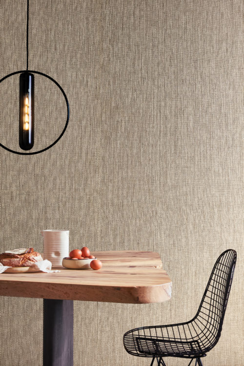 This inviting interior highlights the OAS242 reference, titled Calzada, from Omexco’s Oase collection. The wallcovering, composed of bicoloured paper threads, adds depth and warmth to the space, reflecting a handcrafted aesthetic. The natural contrast of light and dark tones enhances its organic appeal, making it a perfect backdrop for this cozy dining setting. A raw-edge wooden table with a black metal base is paired with a minimalist wireframe chair, evoking a blend of rustic and industrial elements. Above, a striking black circular pendant light provides a bold visual statement, complementing the earthy textures of the space.