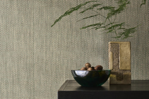 This elegant setting features the OAS243 reference, titled Calzada, from Omexco’s Oase collection. The wallpaper, crafted from bicoloured paper threads, showcases a rich woven texture that exudes natural authenticity. The interplay of soft beige and muted green hues enhances the organic aesthetic, creating a warm and inviting ambiance. The refined setting is complemented by a dark wooden surface, adorned with a deep green glass bowl filled with walnuts and a textured ceramic vase holding delicate greenery. The careful balance of textures and colors results in a harmonious, nature-inspired composition.