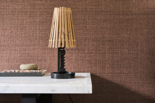 This refined interior highlights the OAS291 reference, titled Cesta, from Omexco’s Oase collection. The wallpaper, composed of delicately woven paper fibres, is dyed with a gentle irregularity, capturing the essence of organic beauty. Its rich, earthy tones exude warmth and a natural elegance, enhancing the overall ambiance. The design is complemented by a striking sculptural table lamp with a bamboo shade, adding an artisanal touch. A marble-topped table with intricate inlaid details serves as a refined surface for books and decorative objects, reinforcing the theme of understated sophistication.