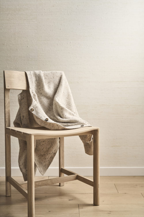 A minimalist and elegant composition showcasing Omexco's Pure collection, reference PU311, a wallpaper crafted from precious silk, revered as the most prized of all natural fibers. The refined texture of the wallcovering exudes understated luxury and adds a natural sophistication to the space. The scene features a wooden chair with clean lines, draped with a textured beige knit cardigan, blending warmth and softness with the raw beauty of the silk wallpaper. The light wooden flooring complements the neutral tones of the wall and furniture, creating a harmonious and serene atmosphere that celebrates natural materials.