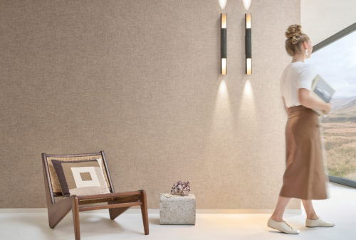 A tranquil setting featuring Omexco's Pure collection, reference PU381, composed of warp and weft weaving of wool yarn in mottled colours. The wallcovering’s nostalgic design adds warmth and texture to the space, creating a cozy yet refined ambiance. The scene includes a mid-century modern wooden lounge chair with a cane backrest and a square-patterned cushion, positioned on a soft white floor. To the left, a stone block displays a striking arrangement of pink and white barnacle shells. On the wall, two minimalist black and gold sconces cast warm, focused light, enhancing the natural tones. To the right, a woman holding a book gazes out of the window at the serene landscape, seamlessly blending the interior with its natural surroundings.