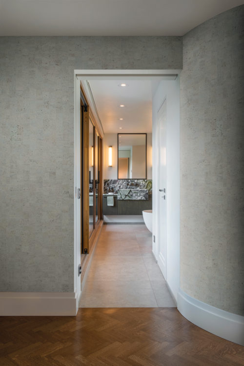 An image of a stylish hallway with a dark herringbone pattern wood floor and a rounded wall on the right. In the background, a bathroom can be seen featuring a freestanding bathtub. On the walls of the hallway, Omexco’s cork wallcovering collection «Casca» is installed, more specifically the design “tactility”, in a blueish grey green colour. This is a faux plain wallcovering, the result of a patchwork of rectangular cork elements which creates a variety of different hues on the wall. The image offers a glimpse into a home that is both visually appealing and comfortable.