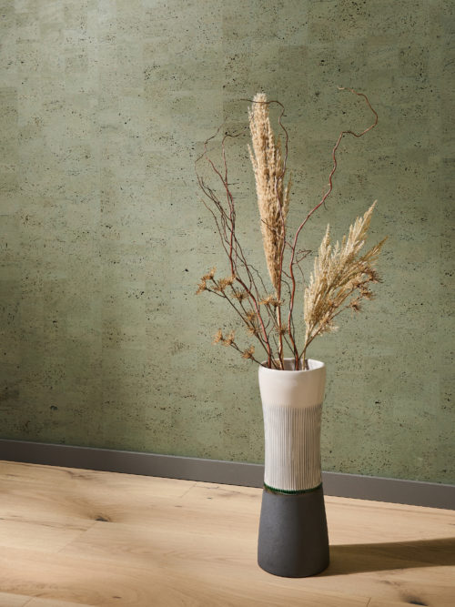 Decor image of a large bicolor ceramic vase standing on an oak wood flooring and holding some dried flowers, twigs and pampas grass. In the background we see Omexco’s cork wallcovering collection “Casca", more specifically the design “tactility” in a moss green colour. This is a faux plain wallcovering, the result of a patchwork of rectangular cork elements which creates a variety of different hues on the wall. The sunlight casts a warm, golden glow on the floor and wall.