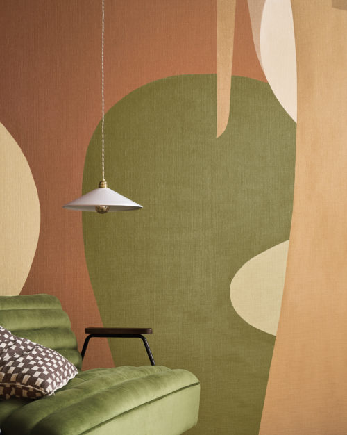 This artistic living space features the OAS211 reference, titled Alegre, from Omexco’s Oase collection. The wallcovering is a printed paperweave panoramic design, inspired by naïve art, capturing the vibrant essence of South American landscapes. Its flowing organic shapes and earthy tones create a warm and inviting atmosphere. The rich green velvet sofa complements the mural’s natural hues, while a geometric-patterned cushion adds a touch of modernity. A minimalist pendant light with a brass detail subtly enhances the scene, providing a soft glow that highlights the texture and depth of the wallcovering.