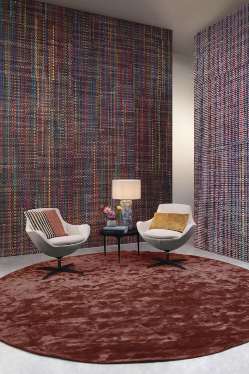From the handweavers loom to your living room. On the walls of this impressive living room we see the "Extravaganza" wallcovering design from Omexco's Atelier collection. Colorful recycled sari silk ribbons are handwoven with Abaca. The cosy nook is also decorated with a round burgundy carpet, side table with tamble lamp, two white lounge chairs with colorful pillows and a small bouquet of fresh cut flowers.