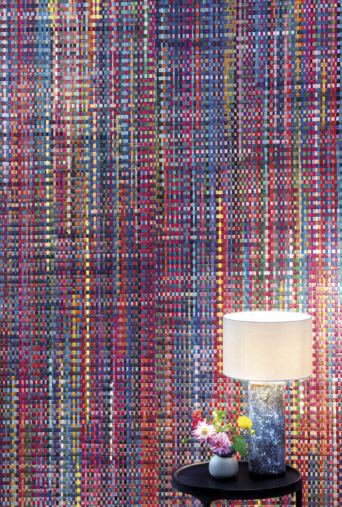 From the handweaver's loom to your living room. Here we see a closeup of the "Extravaganza" wallcovering design from Omexco's Atelier collection. Colorful recycled sari silk ribbons are handwoven with Abaca. In front of the statement wall there is black side table, a table lamp with a beige lampshade and a small bouquet of fresh cut flowers.