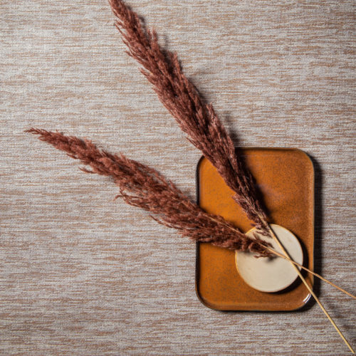 Paper yarn delicately woven into seamless wallcoverings in sand tones. We see a closeup of the wallcoverings as well as decorative feathers and ceramic plates in different shapes.