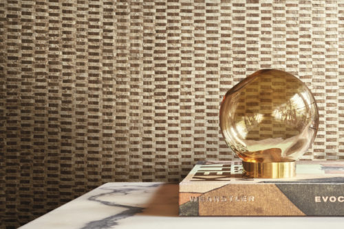 An elegant round glass table lamp, beautifully positioned on top of a book, serves as the focal point in this stylish decor. The backdrop features luxurious capiz wallcoverings from Omexco's Archipelago collection, showcasing a captivating blend of gold and brown shades, creating a sophisticated and inviting atmosphere.