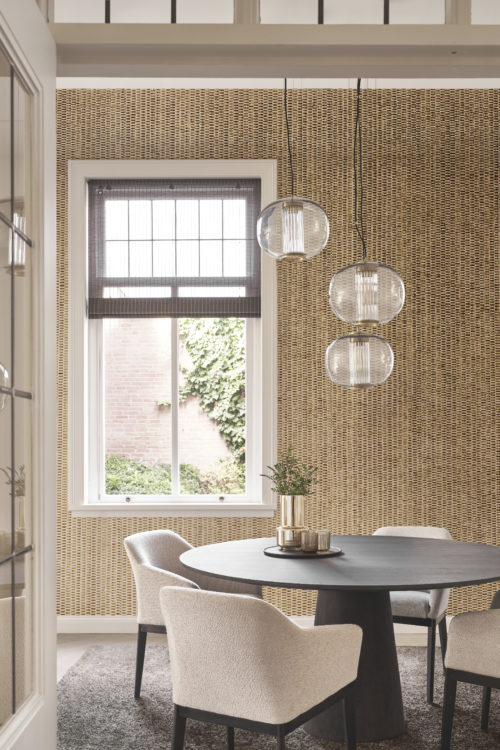 A charming dining room setting featuring a round black wooden table surrounded by four comfortable, soft beige chairs. Above the table, a hanging lamp with three graceful round lampshades illuminates the space. A cozy grey fluffy carpet enhances the dining area's warmth and comfort. The backdrop showcases Omexco's capiz mother of pearl wallcoverings from the Archipelago collection, exhibiting shimmering shades of gold and brown. The textured wallcoverings add a touch of opulence and visual interest to the overall dining room ambiance.