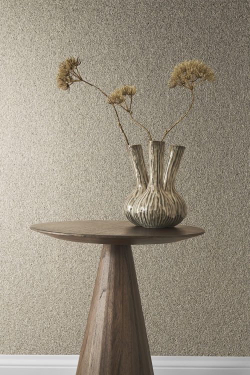 A charming decor composition is captured in this photo, featuring a wooden side table adorned with a distinctive fennel-shaped vase. Dried flowers grace the vase, adding a rustic touch. The backdrop showcases the elegance of Omexco's Archipelago collection, featuring embossed grey mica wallcoverings that contribute to the overall sophistication of the scene, creating a harmonious blend of natural elements and contemporary design.