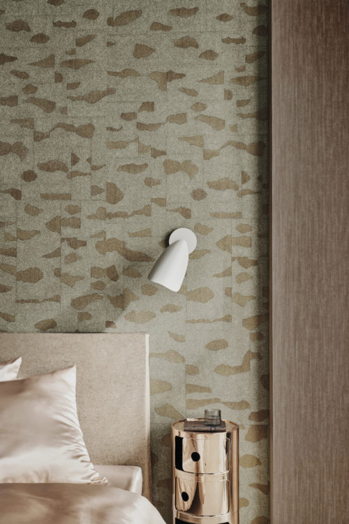 This close-up of a chic bedroom showcases an inviting bed adorned with champagne-colored sheets, accompanied by a luxurious gold metal nightstand and a sleek white wall lamp. The backdrop features fine stardust Omexco wallcoverings, where mica is delicately printed with an organic design, evoking a sense of nature. The combination of soft hues, metallic accents, and nature-inspired textures creates a serene and sophisticated atmosphere in this stylish bedroom setting.