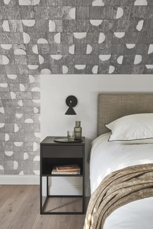 A chic bedroom ensemble captured in this photo includes a modern bed, complemented by a sleek black nightstand and a matching wall lamp. The backdrop showcases the exquisite Omexco wallcovering collection, Archipelago, featuring a stylish grey patchwork adorned with elegant white capiz shells. The combination creates a sophisticated and contemporary ambiance, blending seamlessly for a visually appealing decor.