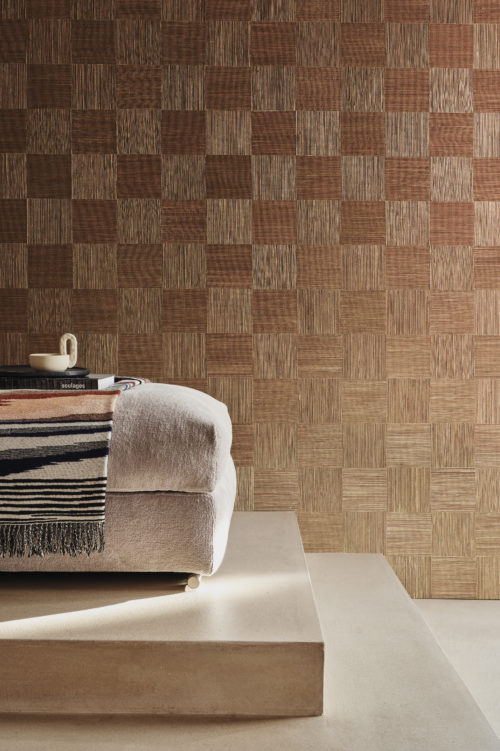 Elegant decor featuring a beige floor with varied steps, adorned with an inviting beige sofa adorned with books, a vibrant blanket, and a ceramic cup. The backdrop showcases Omexco's handmade natural wallcovering collection, Intuition, featuring an intricate patchwork of cattail in burgundy. The natural fiber is meticulously woven and hand-painted, exuding artisanal allure. Illuminated by soft daylight, the scene radiates warmth and sophistication.