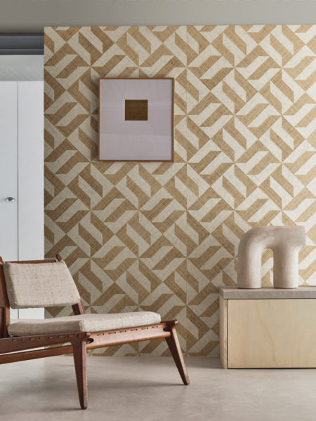 A captivating decor shot featuring an ethnic Scandinavian-style wooden chair on the left and a light wood cupboard on the right adorned with a ceramic decorative element. The grey cast floor provides a neutral foundation, while the backdrop showcases Omexco's Intuition wallcoverings—a raffia patchwork displaying a striking geometric design with yellow and white shapes, creating a harmonious and stylish interior composition.