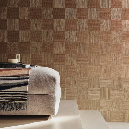 Elegant decor featuring a beige floor with varied steps, adorned with an inviting beige sofa adorned with books, a vibrant blanket, and a ceramic cup. The backdrop showcases Omexco's handmade natural wallcovering collection, Intuition, featuring an intricate patchwork of cattail in burgundy. The natural fiber is meticulously woven and hand-painted, exuding artisanal allure. Illuminated by soft daylight, the scene radiates warmth and sophistication.