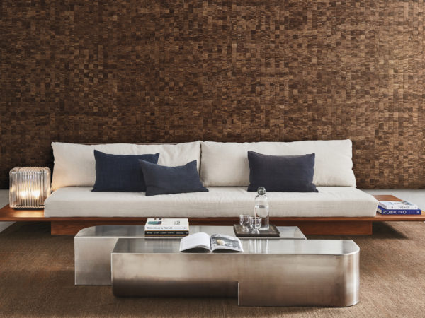 Stylish living room boasting a gray cast floor and a plush brown carpet. The centerpiece is a large minimalist sofa with a wooden frame, adorned with light beige cushions and dark blue pillows. Flanking the sofa are glass table lamps and piles of books on each side. In front of the sofa, a silver-colored metal coffee table displays more books. The true focal point is the striking wall adorned with Omexco's handmade natural "Intuition" wallcoverings. The mosaic-like mulberry weave in shades of brown adds a captivating and textured backdrop, enhancing the room's sophistication.