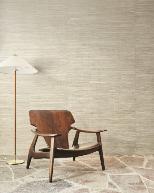 The focal point of this elegant space is the exquisite blend of natural elements and mid-century modern design. Beneath our feet lies a captivating natural stone floor, its cool, earthy tones grounding the room with timeless beauty.  Taking center stage is a sleek mid-century modern wooden armchair, its clean lines and organic curves inviting relaxation and contemplation. Positioned beside it, a slender floor lamp casts a warm glow, its beige lampshade adding a soft touch of sophistication to the ambiance.  As we gaze upon the backdrop, we're drawn to the captivating allure of Omexco's handmade natural wallcoverings collection "Intuition". A mesmerizing textured cattail weave adorns the walls, enveloping the space in a serene palette of shades of beige.