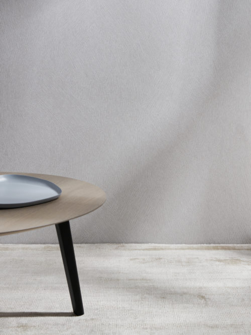 An image of a wooden side table with a natural and earthy feel. A beige carpeted floor creates a neutral base while a neutral Omexco wallcovering with a brushed chalk effect serves as the backdrop. These High Performance Patina wallcoverings meet the essential technical standards while being eco-conscious. On top of the side table an organic shaped plate is placed, adding texture and visual interest to the scene.