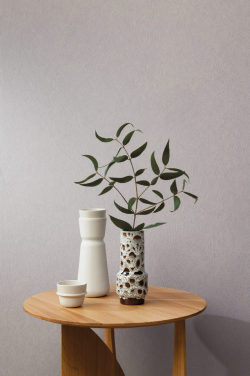 Closeup on a mini decor with a wooden side table and three organic shaped vases placed on top of it. One of the vases holds a small branch with leaves. On the wall we see the High Performance Patina wallcovering range by Omexco. This particular design, printed in light grey, is inspired by a special painting technique that creates a brushed chalk effect on the wall.