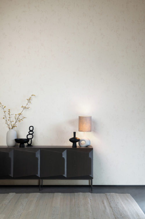 A contemporary, rather minimalist room with clean lines and a restrained palette. A black console cabinet is placed against the wall. Decorative elements such as black and white organic shaped vases and a table lamp are placed on top of it. A soft, light grey rug lays on the concrete floor. On the walls we see Omexco’s High Performance Patina wallcoverings in off white. The design is Inspired by the structure of a sandstone. Very organic.