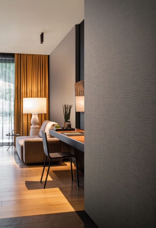 This image shows a hotel room with orange curtains, a floor lamp, a two seater sofa and a desk with chair. On the walls we see Omexco's High Performance Patina wallcoverings, a printed non-woven collection created for residential, commercial and hospitality interiors all over the world. The design, a print of vertical stripes combined with horizontal lines, is called 'Groove' and shown here in shades of brown.