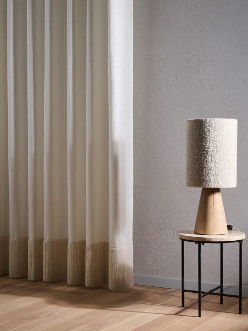 An image of airy and contemporary living room corner with on the right a wooden round side table and a wooden table lamp on top of it. We see a white linen curtain on the left. A wooden parquet floor creates a natural base while a neutral Omexco wallcovering with a brushed chalk effect serves as the backdrop. These High Performance Patina wallcoverings meet the essential technical standards while being eco-conscious.