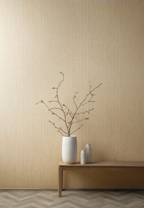 Image of a wooden coffee table with three white organic shaped ceramic vases placed on top of it. One of the vases holds a branch with light pink flower buds. A sold oak herringbone parquet on the floor. On the wall we see the High Performance Patina wallcovering range by Omexco. This particular design, printed in a yellowish beige colour, is very textured and is inspired by the structure of burnt timber, a very popular look nowadays.