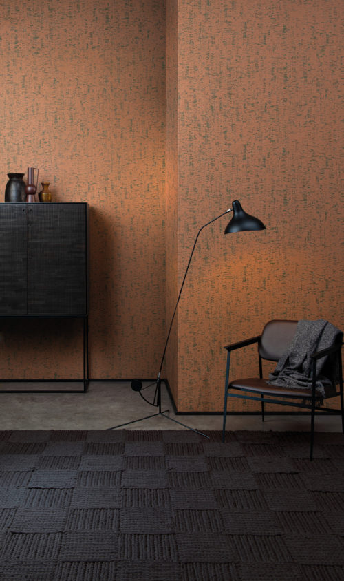 Contemporary interior with a black wooden cupboard, a minimalistic black floor lamp and a comfortable chair with black metal frame. The walls show printed non-woven wallcoverings by Omexco. The design, shown here in burnt orange, was inspired by the clay walls and an old painting technique in which thin lime plaster is spread on the wall with a  block brush.