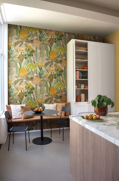Example of a modern kitchen with flowery botanical wallcoverings and a cosy breakfast nook next to the window. Shown here are Omexco’s exuberant “Birds of Paradise” wallcoverings, a fabulous foliage in shades of yellow, lime green, khaki and beige. The inviting kitchen also has a kitchen island with a marble tablet and a large white pantry.