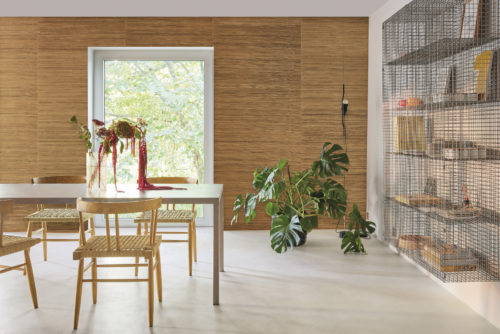 This image depicts a modern and bright living room witha grey cast floor. On the left side of the room, a minimalist metal table is surrounded by rattan chairs, creating a chic and inviting seating area. A bouquet of flowers adorns the table. Behind the table, a large window offers a view of a tree, inviting natural light to illuminate the room. On the right side of the image, a Monstera plant adds a touch of greenery, while a metal grid bookcase stands tall. In the background, Omexco’s natural handmade Intuition wallcoverings serve as a backdrop. The design features a minimalist weave in orange-brown, highlighting the inherent beauty of youthful abaca skin fibre.