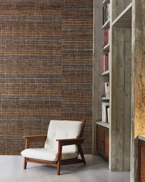 This image depicts a cozy reading corner nestled within a loft-style living room with high walls and a grey cast floor. In the corner, a wooden mid-century modern armchair with a white cushion invites relaxation and contemplation. Next to the armchair stands a tall open bookcase, its built-in shelves displaying a collection of literary treasures waiting to be explored. The focal point of the room is Omexco’s handmade natural wallcoverings collection “Intuition”. The wallcovering design reveals itself in shades of brown that intertwine harmoniously, punctuated by the skillful weaving of various materials such as bakbak, bacnoc, and paper yarn. This eye-catching wallcovering adds depth and texture to the space, infusing it with a sense of warmth and sophistication.
